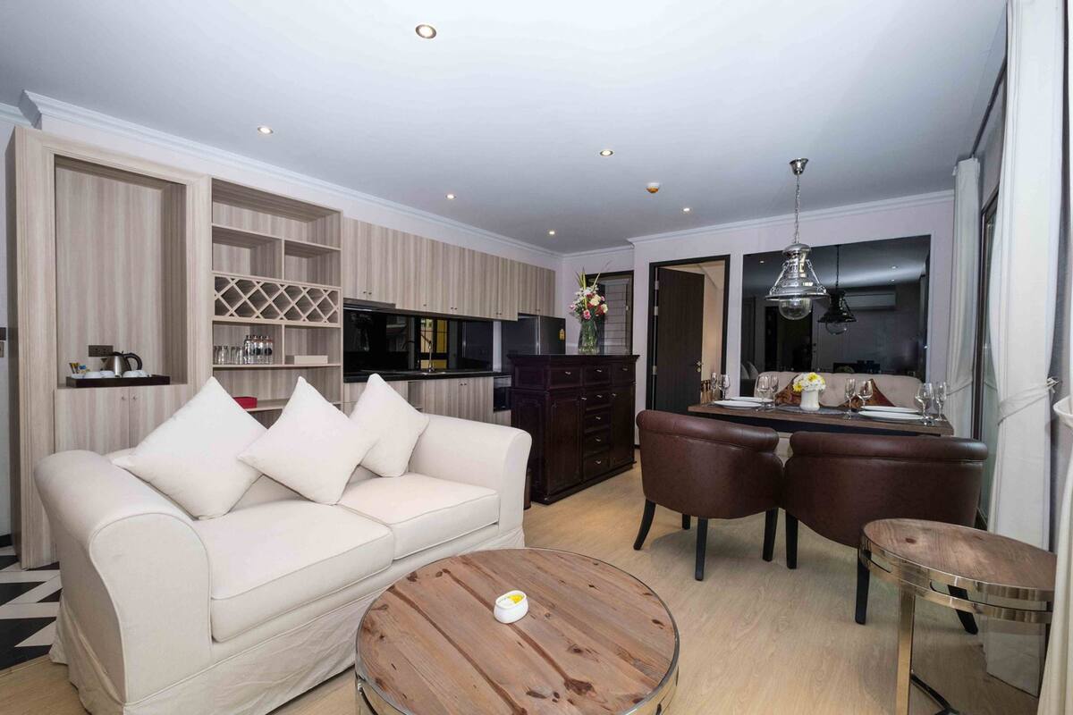 A modern living room with a sofa, coffee table, dining table, and chairs. There is a kitchen area with cabinets and a refrigerator.