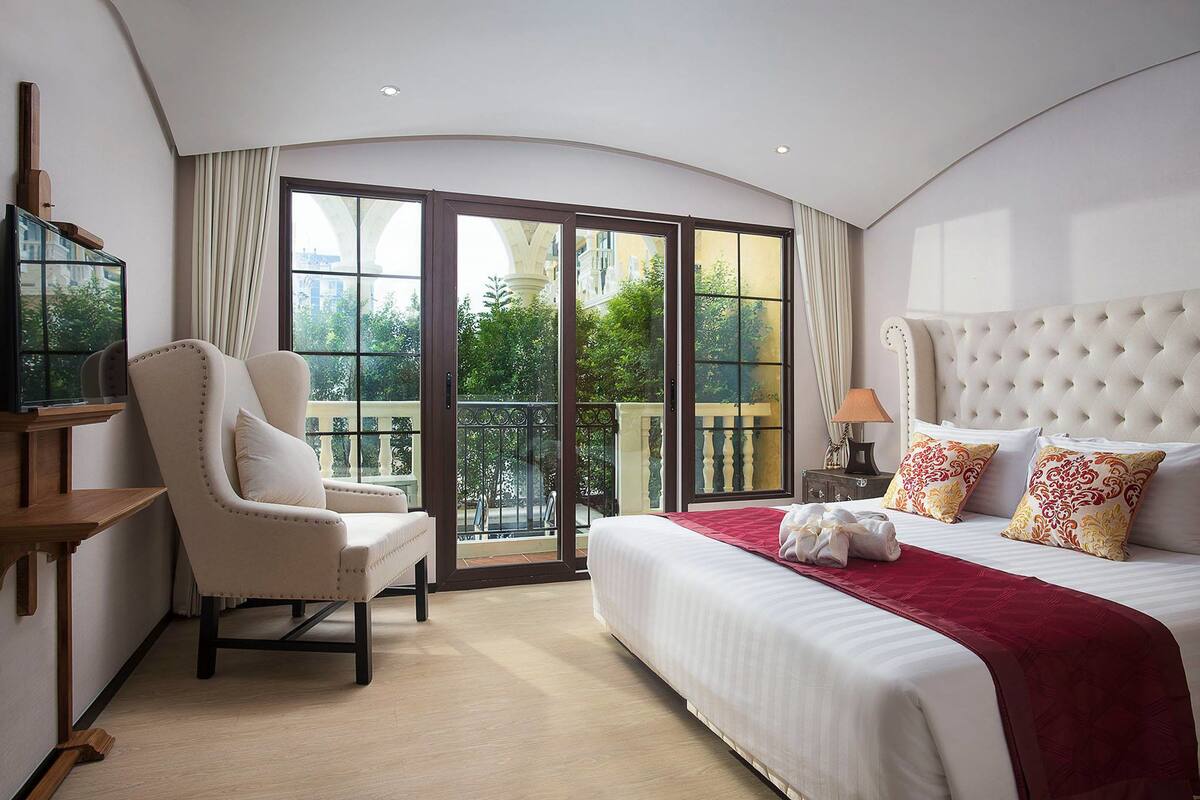 A hotel room with a large bed, a chair, a TV, and a balcony with a view of greenery.