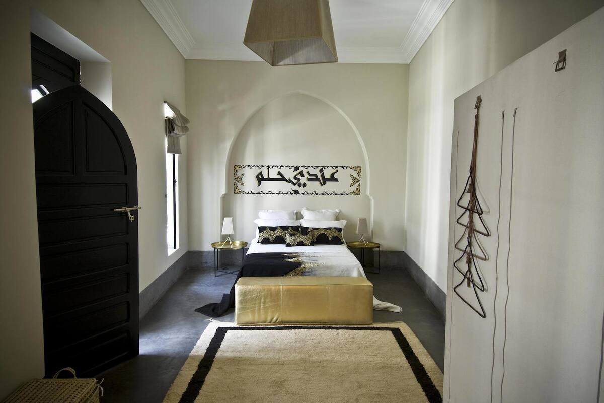A bedroom with a large bed, a nightstand, a lamp, a wall-mounted artwork, and a black door.