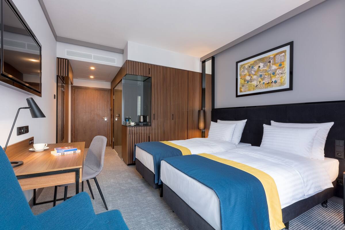 A hotel room with two beds, a desk with a chair, a coffee maker, and a framed artwork on the wall.
