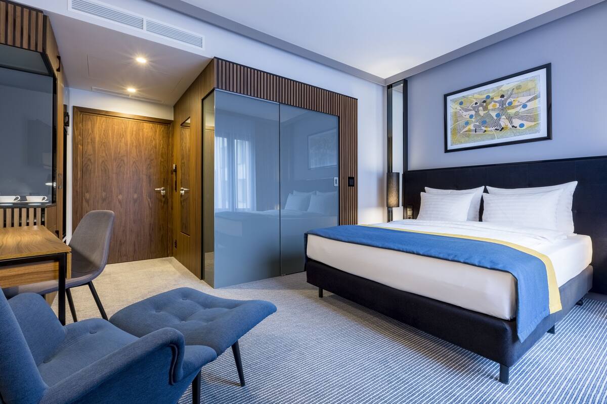 A modern hotel room with a bed, a desk, a chair, a wardrobe, and a painting on the wall.