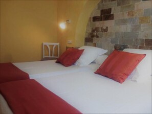 A hotel room with two beds, each with white linens and red pillows. The room has a stone wall and a yellow wall. - Hotel La Casa del Abuelo (Tapia de Casariego)