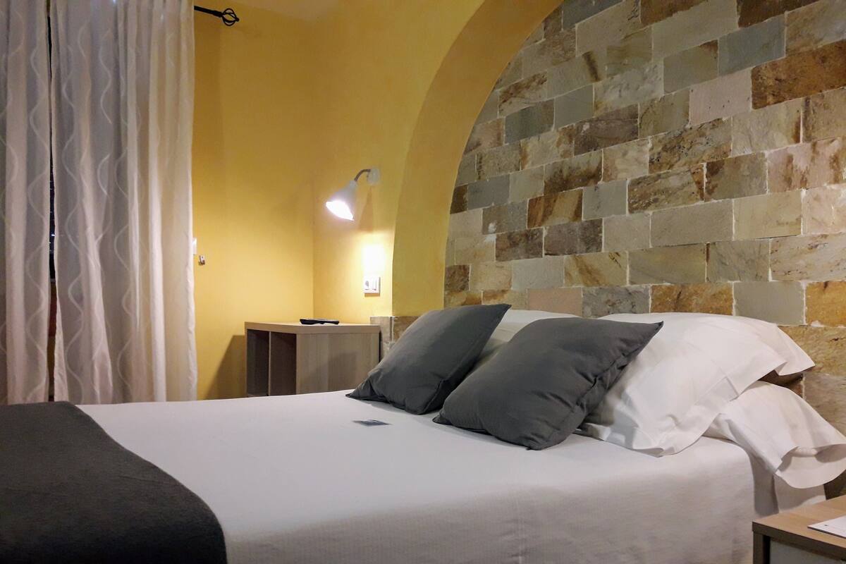 Ein Bett mit weißen Leinen und zwei Kissen, ein Nachttisch, eine Lampe sowie eine Steinwand.