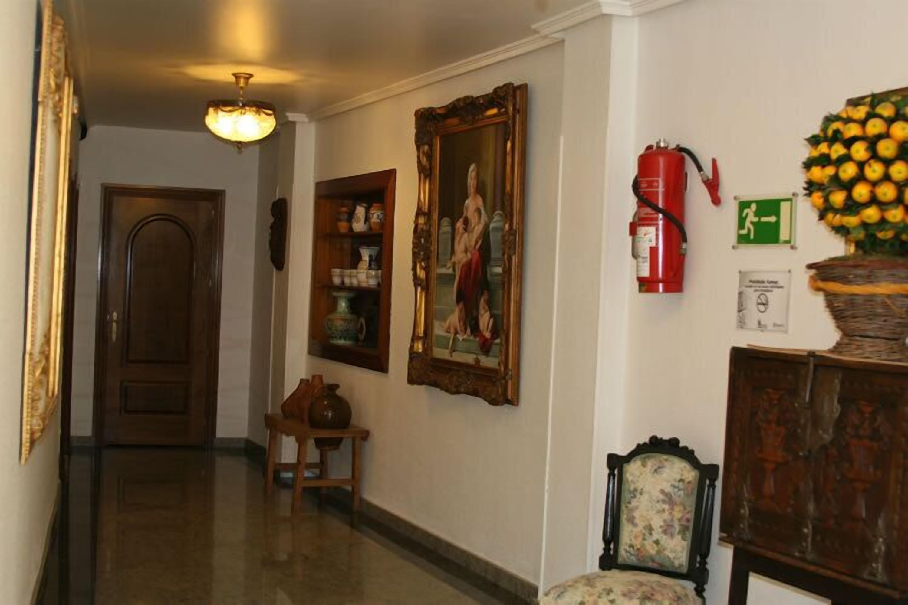 A hallway with a wooden door, a small table with a vase, a chair, a wall-mounted painting, a fire extinguisher, and a wall-mounted sign.