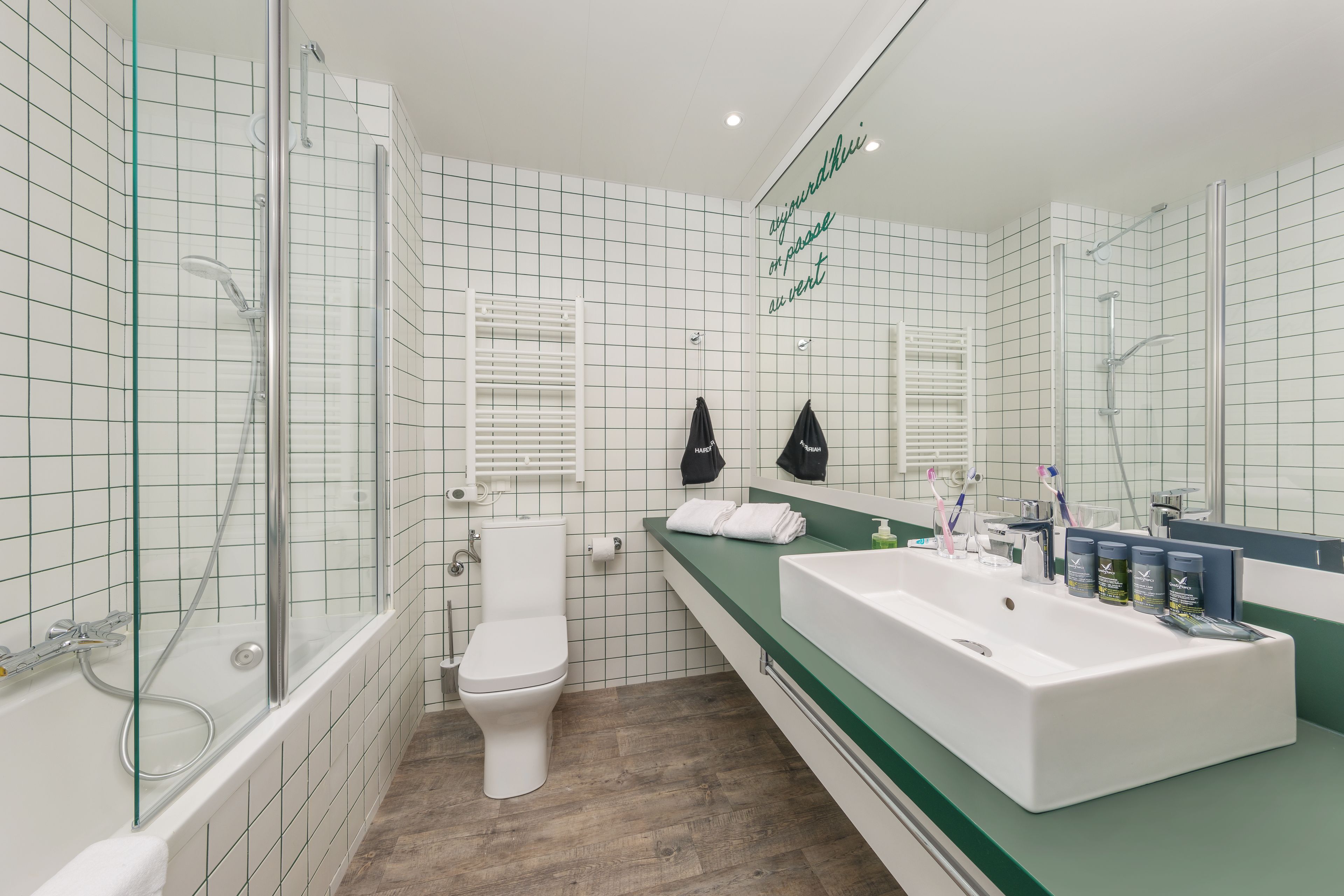A modern bathroom with a glass-enclosed shower, a white toilet, a large sink with a mirror, and a green countertop.