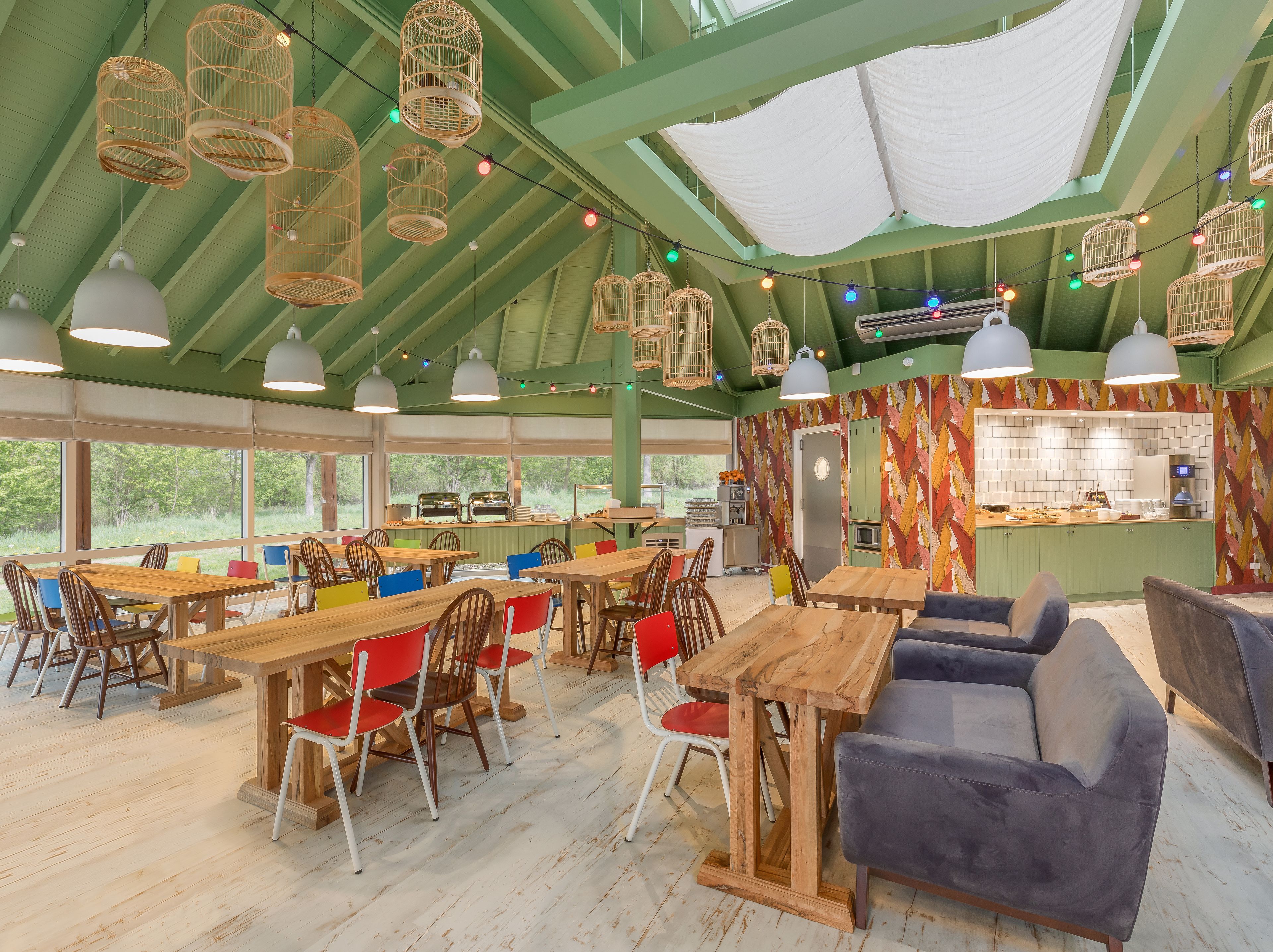 A rustic interior with wooden tables and chairs, a kitchenette, and large windows offering a view of the outdoors.