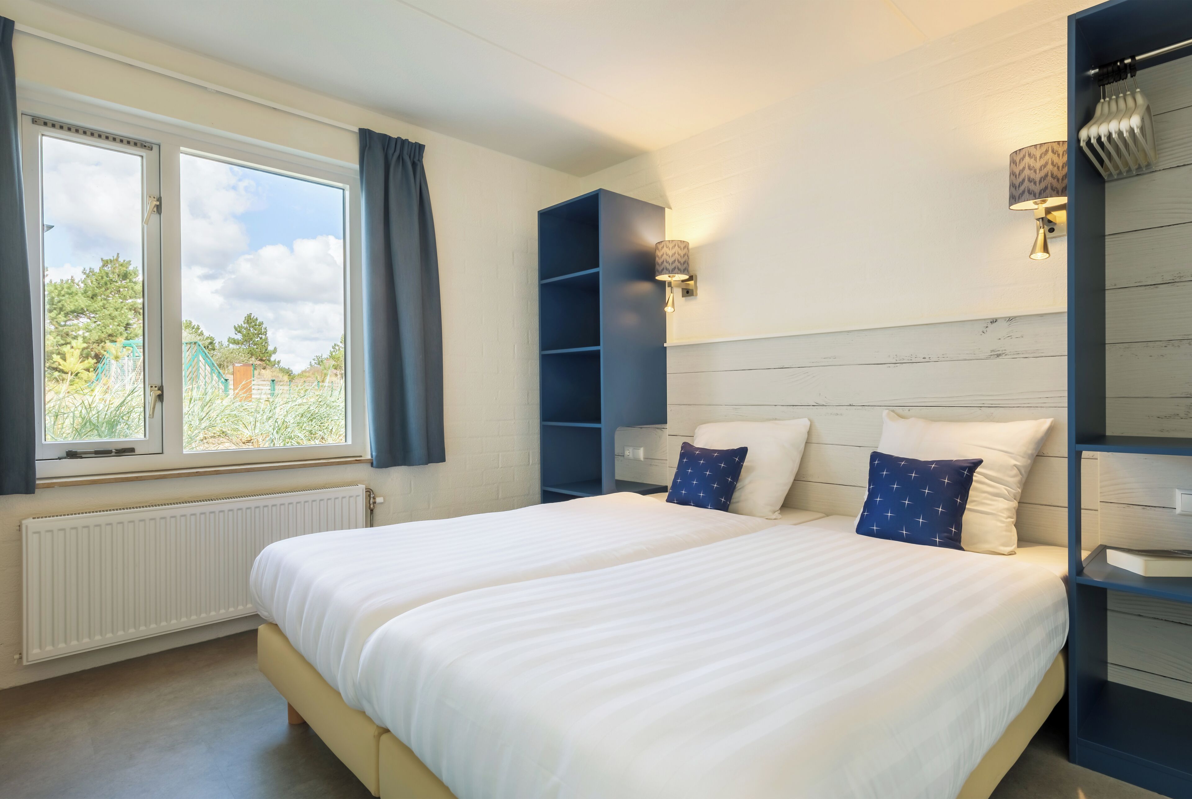 A bedroom with a large bed, a window with curtains, a built-in bookshelf, and a wall-mounted lamp.