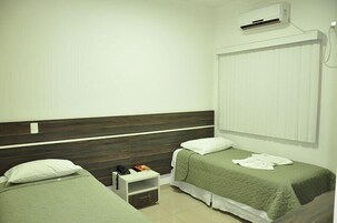 Una habitación pequeña y limpia con dos camas individuales, una mesita de noche con teléfono y libro, y un equipo de aire acondicionado en la pared.