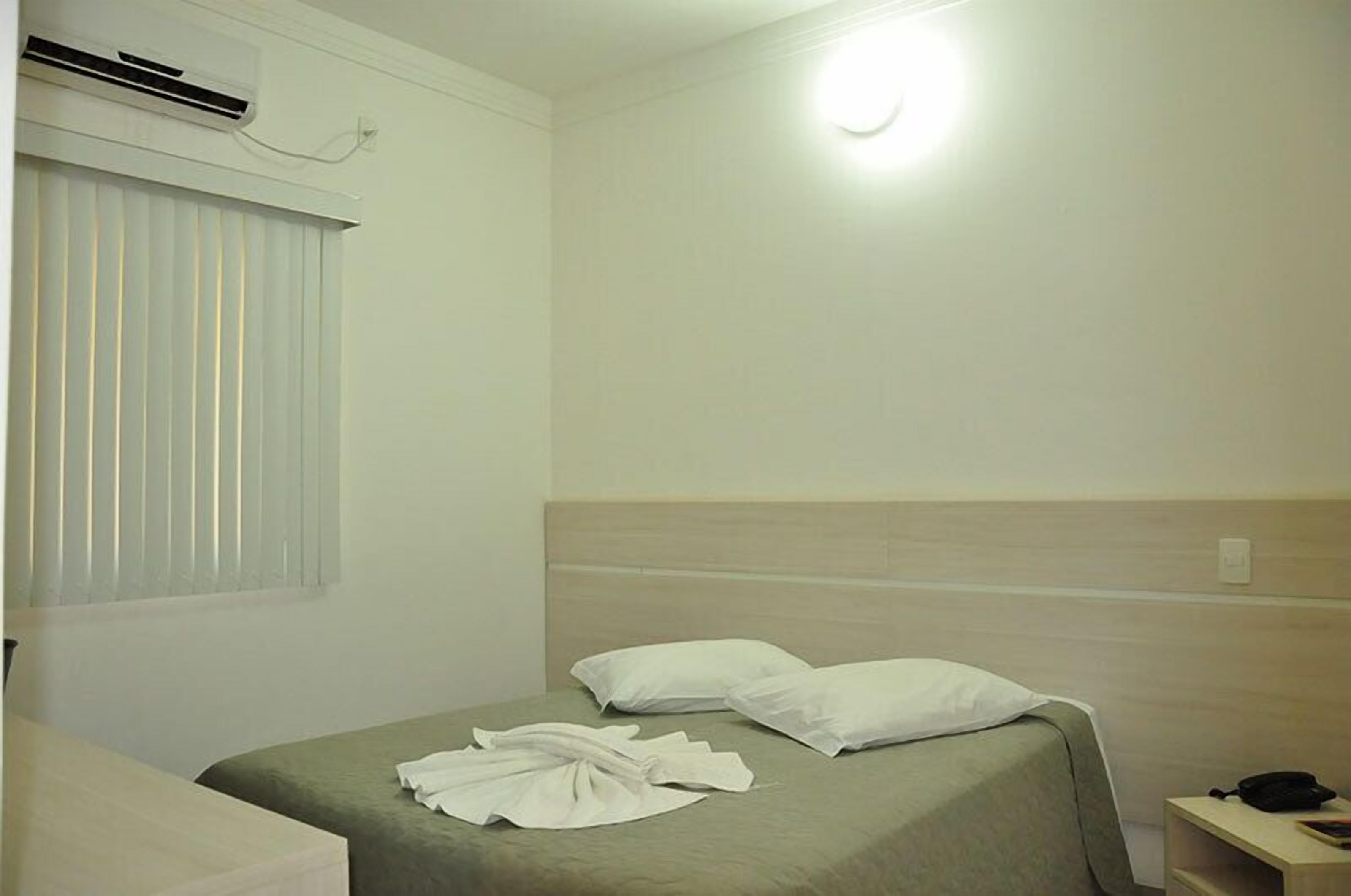 A hotel room with a single bed, a bedside table, a lamp, a window with blinds, and an air conditioning unit.
