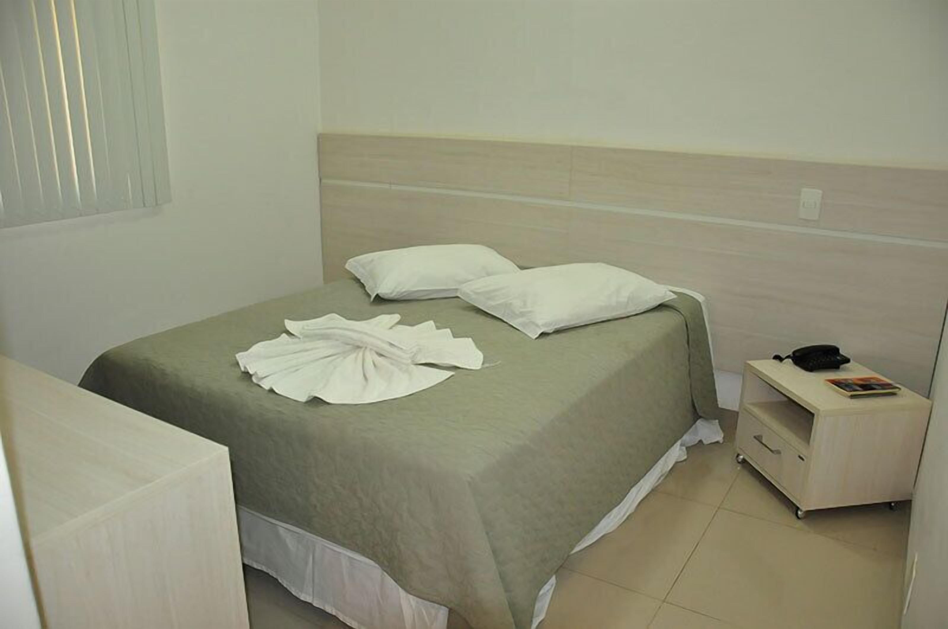A neatly made bed with a green bedspread and white pillows, a bedside table with a phone and a magazine, and a window with blinds.
