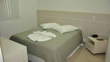 A neatly made bed with a green bedspread and white pillows, a bedside table with a phone and a magazine, and a window with blinds.