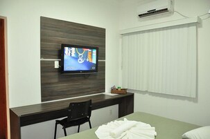 Habitación de hotel con televisión de pantalla plana montada en una pared de paneles de madera, un escritorio con silla, una cama con ropa de cama blanca y una ventana con persianas.
