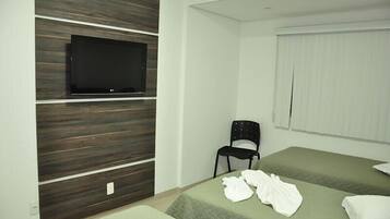 Habitación de hotel con televisor de pantalla plana montado en una pared de paneles de madera, una silla negra, dos camas con ropa de cama verde y una ventana con persianas.