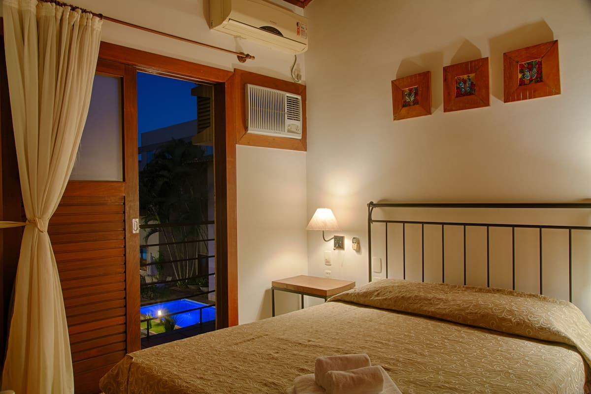 Een hotelkamer met een bed, een tafeltje, een nachtkastje, een lamp en uitzicht op een zwembad.