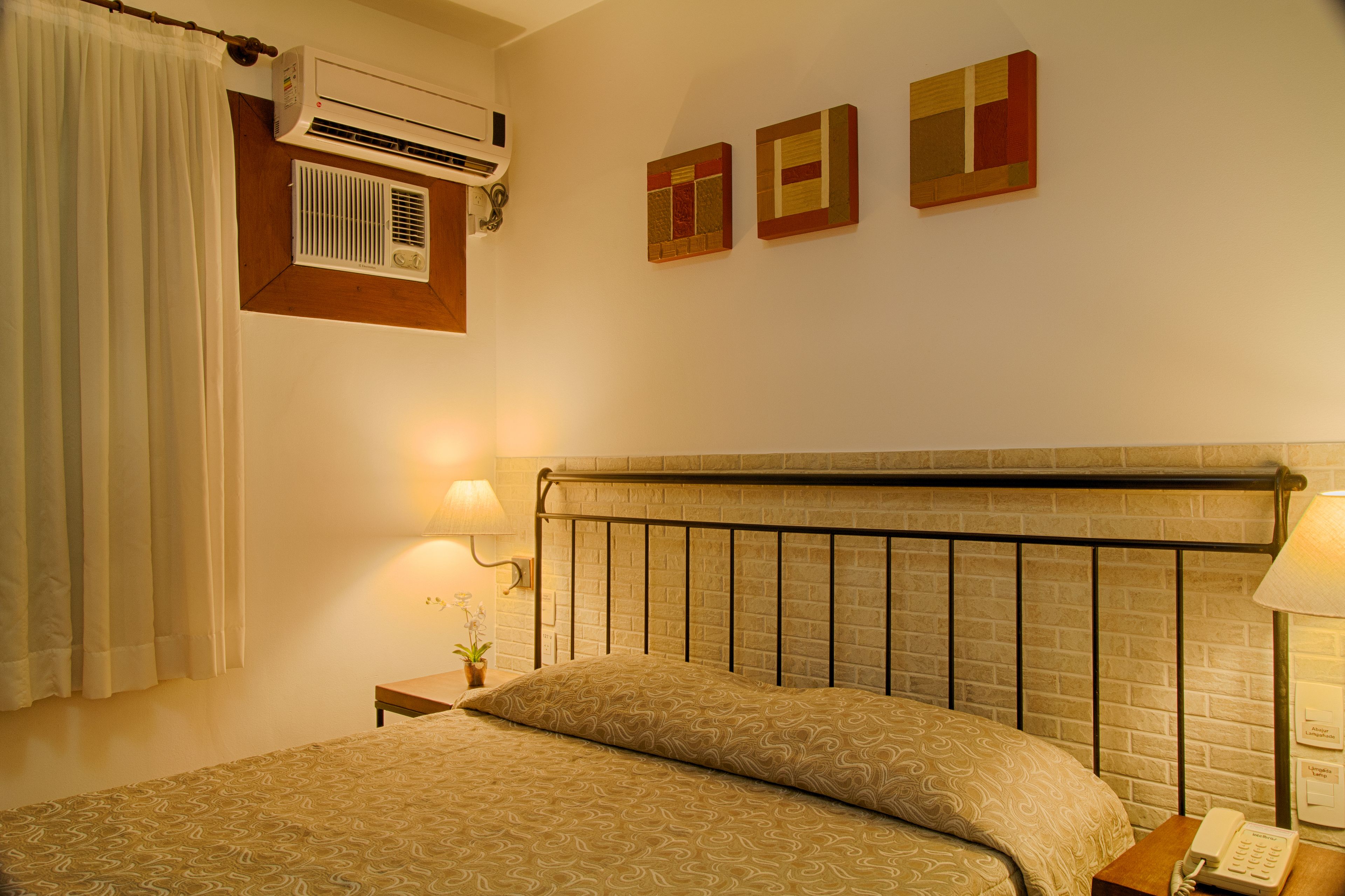 Una habitación de hotel con cama, mesita de noche, lámpara, aire acondicionado y dos obras de arte colgadas en la pared.