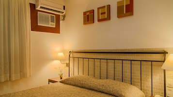 Una habitación de hotel con cama, mesita de noche, lámpara, aire acondicionado y dos obras de arte colgadas en la pared.