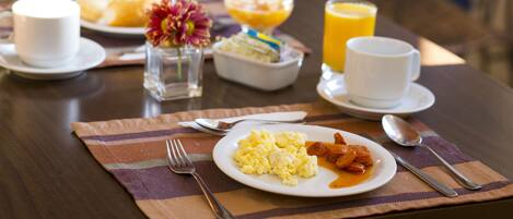 A breakfast table with plates of scrambled eggs, bacon, and toast, a bowl of fruit, a glass of orange juice, and a cup of coffee.