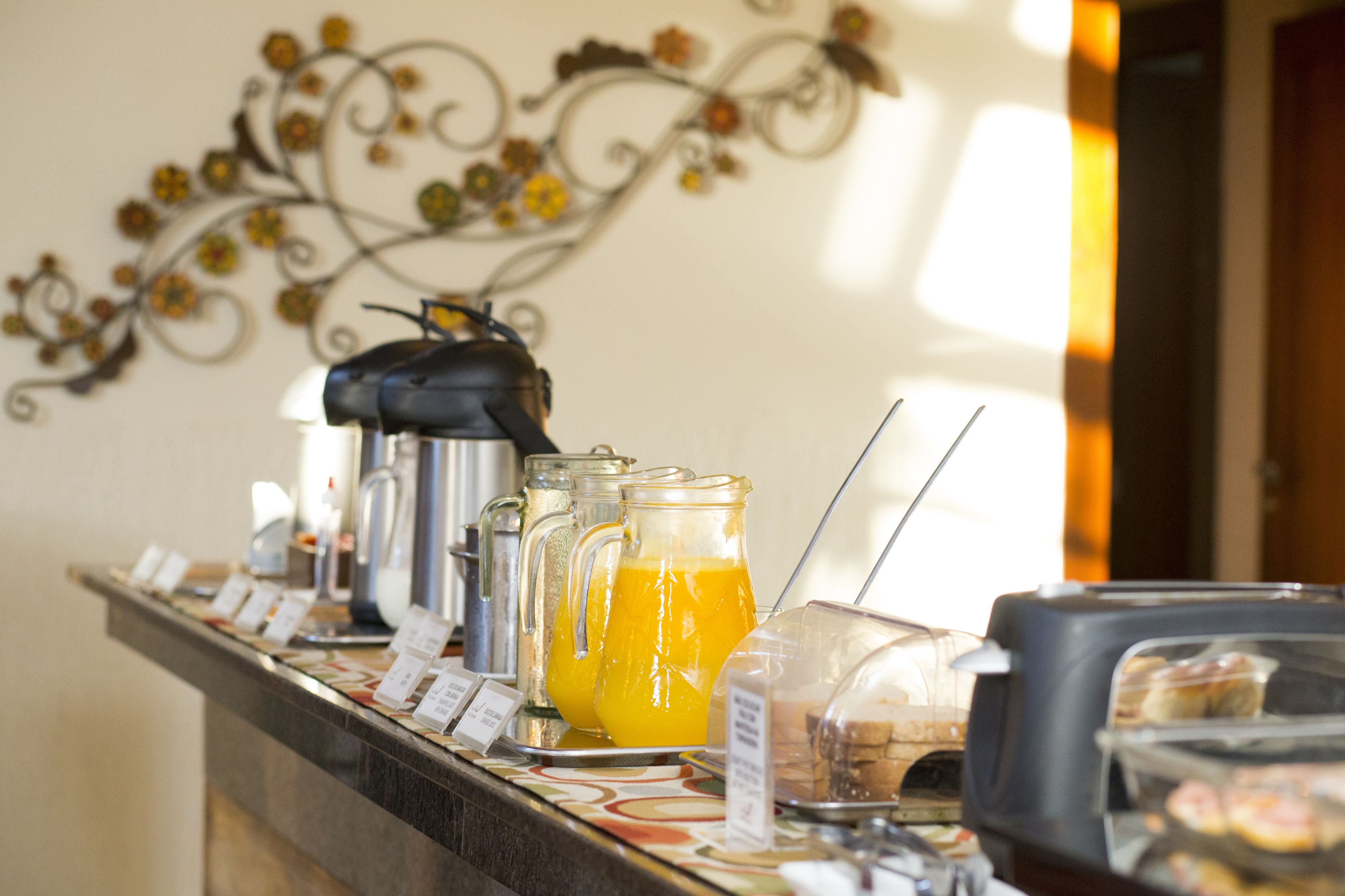 A buffet counter with various food items and beverages, including a pitcher of orange juice, a coffee machine, and a container of pastries.