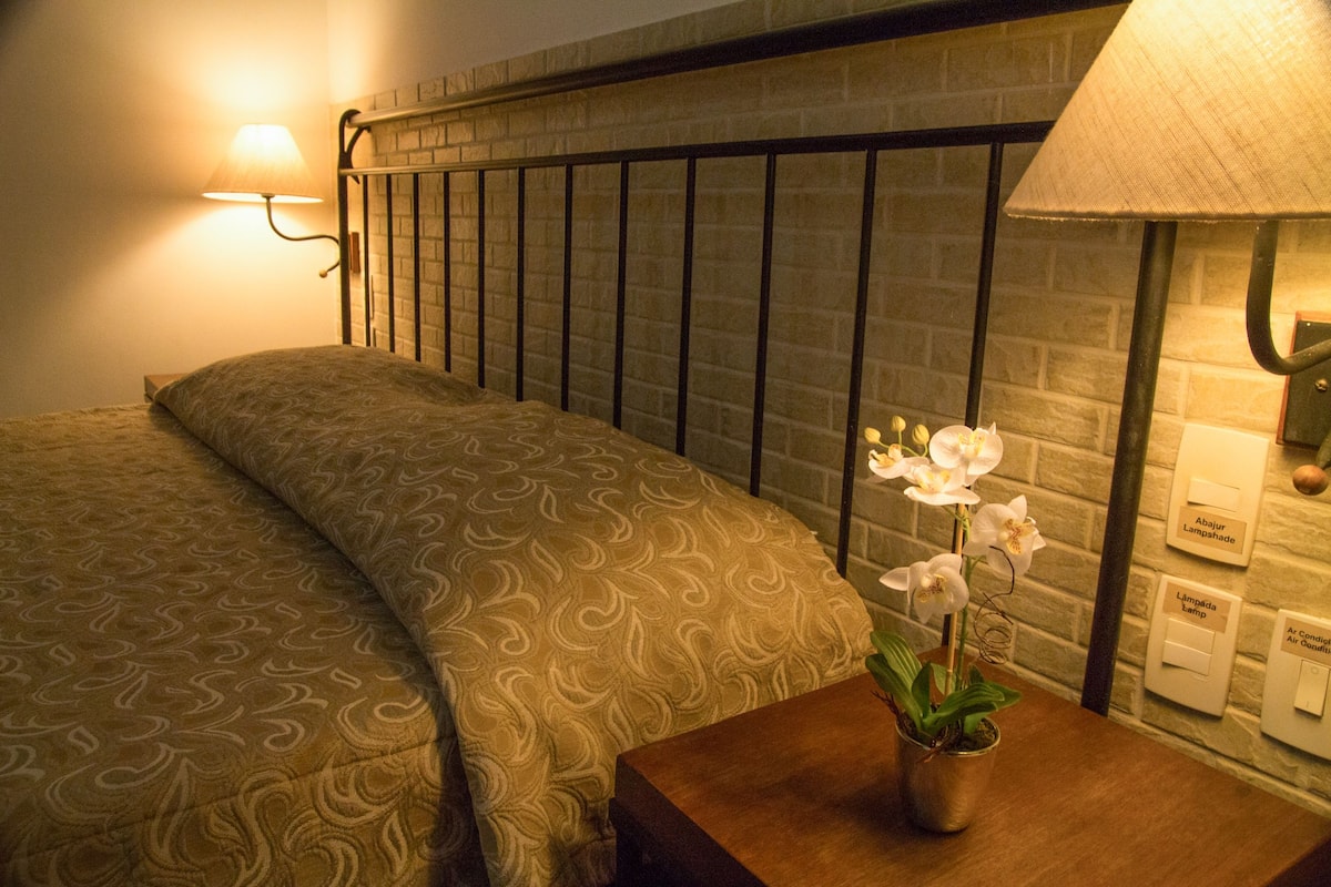 Een net opgemaakt bed met een bedsprei met een patroon, een nachtkastje met een lamp en een potorkidee, en een hoofdbord met een baksteenmotief.