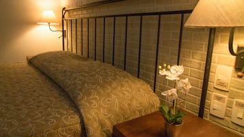 Una cama bien hecha con una colcha estampada, una mesita de noche con una lámpara y una orquídea en maceta, y un cabecero con diseño de ladrillo.
