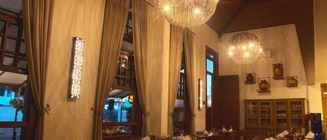 A restaurant with multiple dining tables and chairs, large windows with curtains, and decorative lighting fixtures.