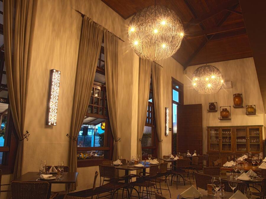 A restaurant with multiple dining tables and chairs, large windows with curtains, and decorative lighting fixtures.