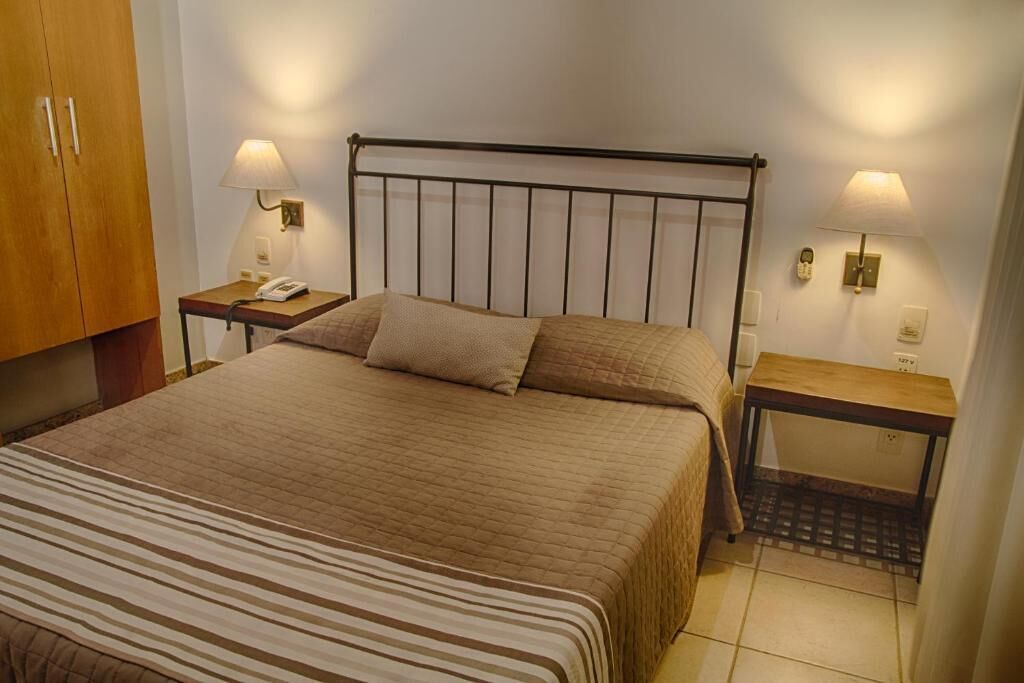 Een net opgemaakt bed met een gestreept sprei, twee nachtkastjes met lampen en een houten garderobe.