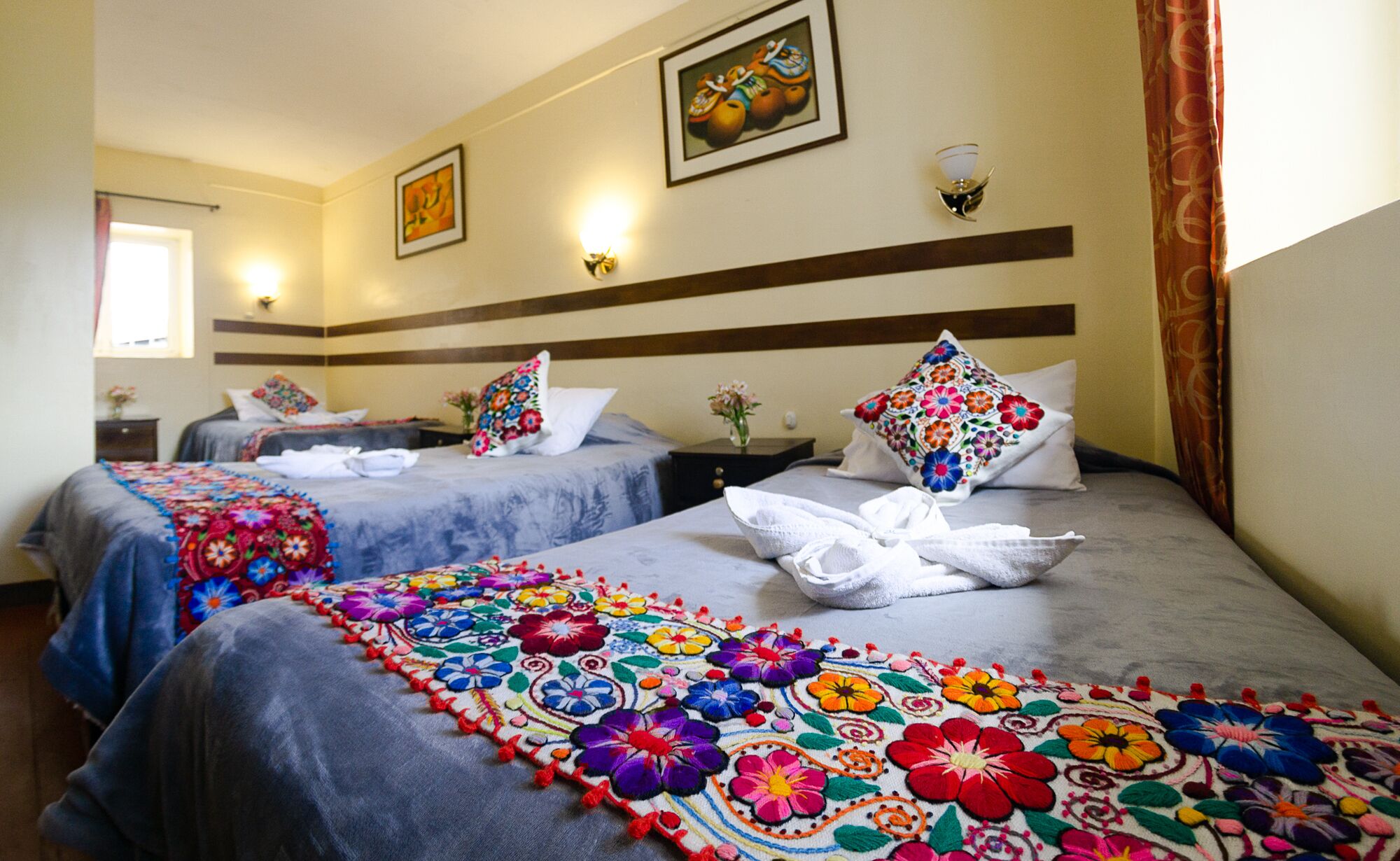 A hotel room with two beds, each with a colorful floral bedspread, a nightstand with a vase of flowers, and a painting on the wall.