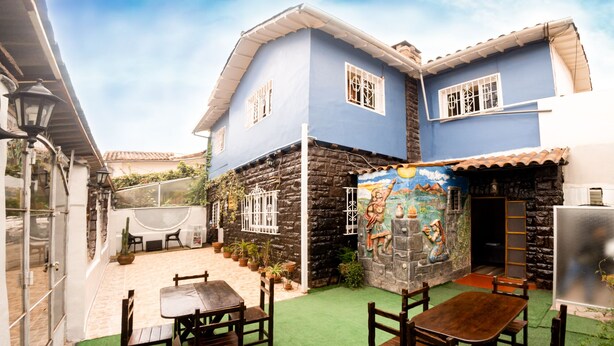 A courtyard with a blue building, stone walls, and a mural. There are wooden tables and chairs. - Samay Inka Inn (Cusco)