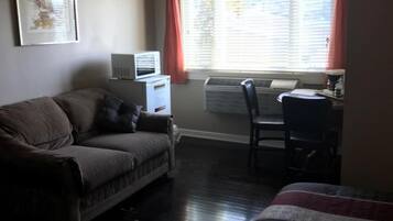 A room with a dark wood floor, a gray sofa with a cushion, a bed with a patterned blanket, a desk with a chair, a window with red curtains, and a framed picture on the wall.