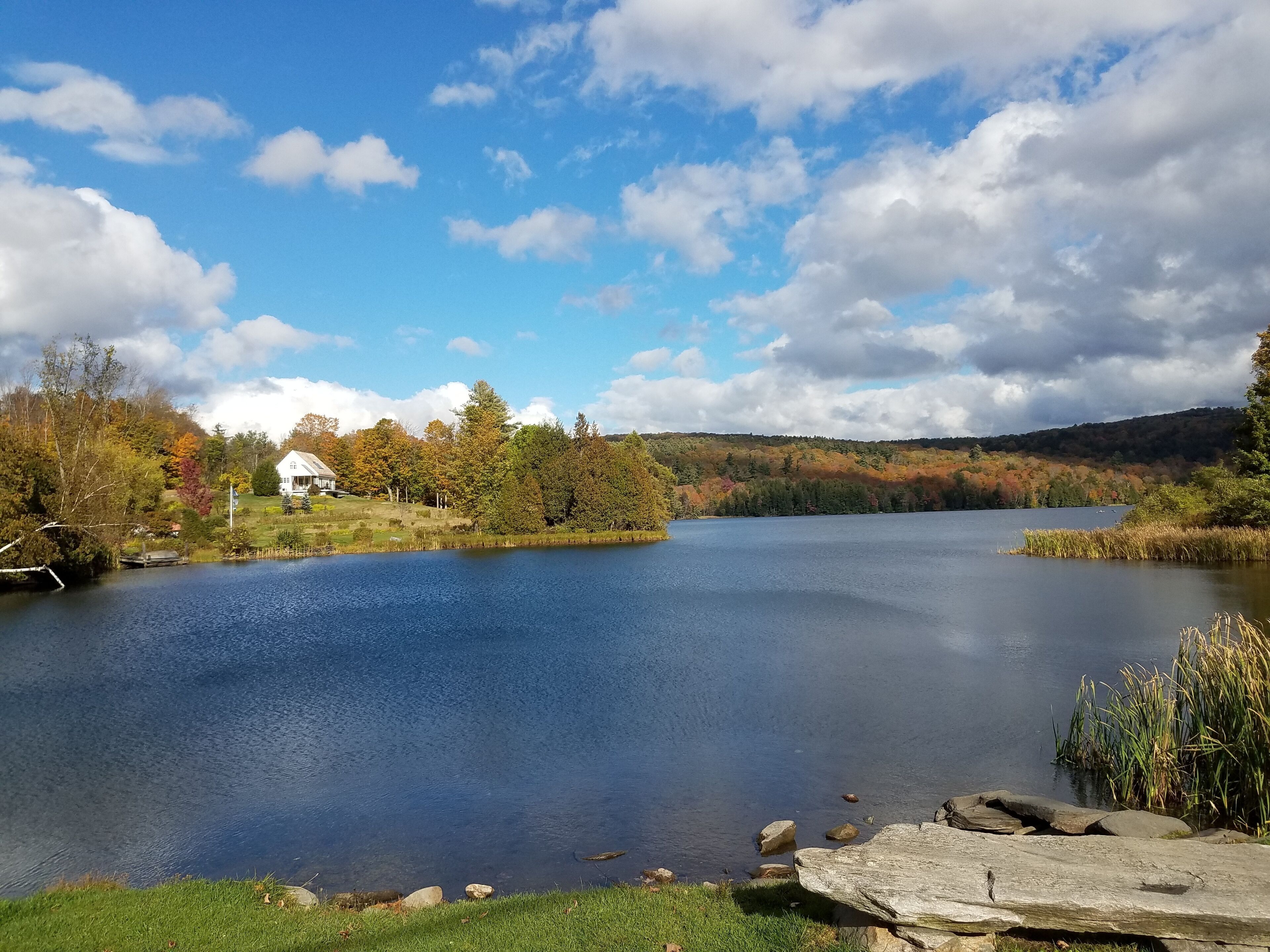Un lago tranquilo con una casa en la orilla, rodeado de árboles de colores otoñales y un cielo despejado.