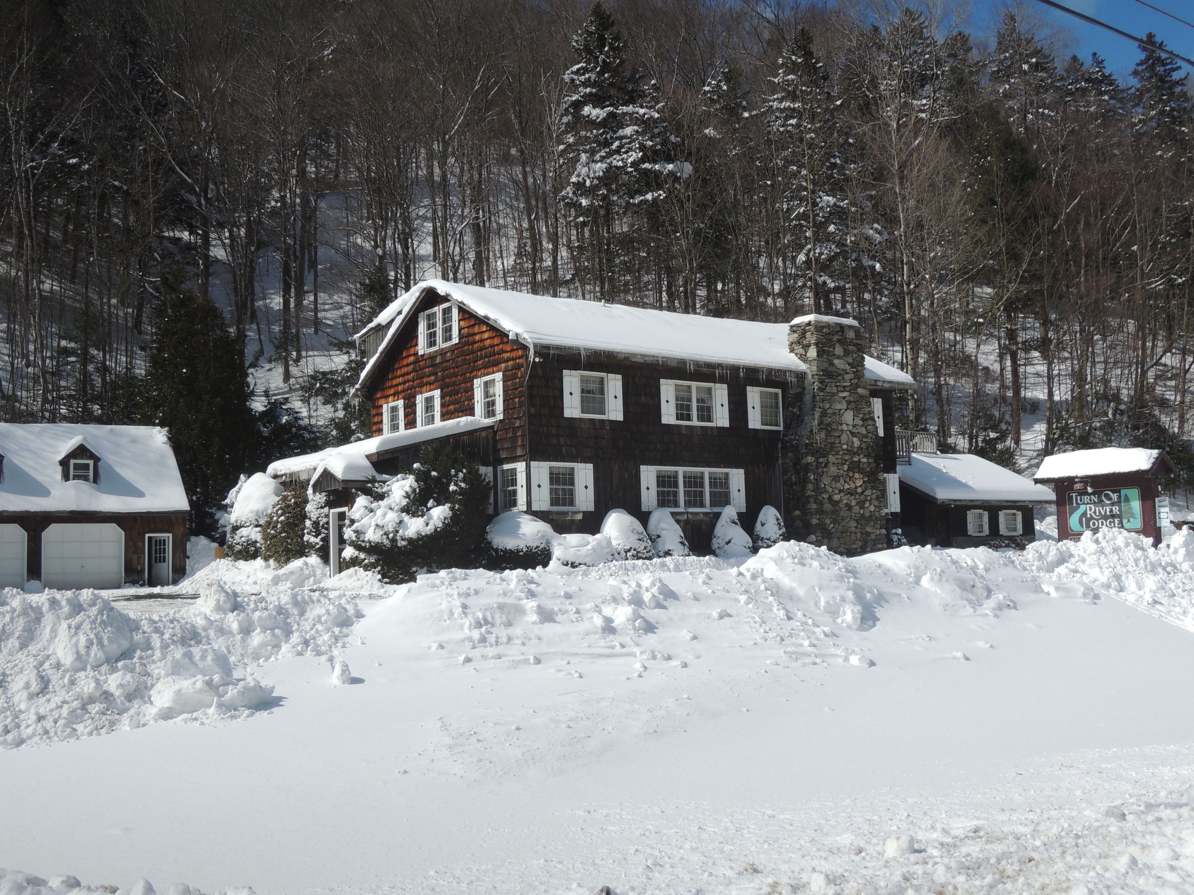 Eine rustikale Lodge mit einem Steinkamin, umgeben von schneebedeckten Bäumen und wolkenlosem Himmel.