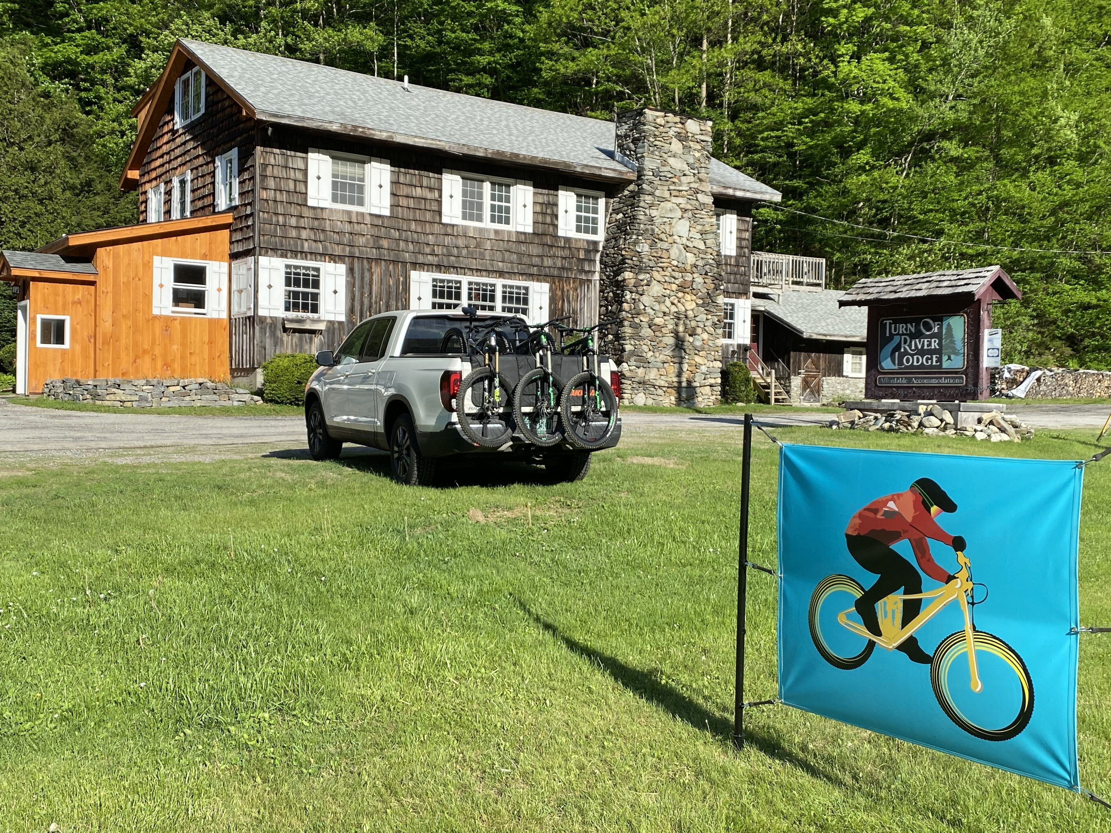 Un chalet rustique avec une cheminée en pierre, une enseigne indiquant « Turn of River Lodge », et un camion garé avec des vélos.