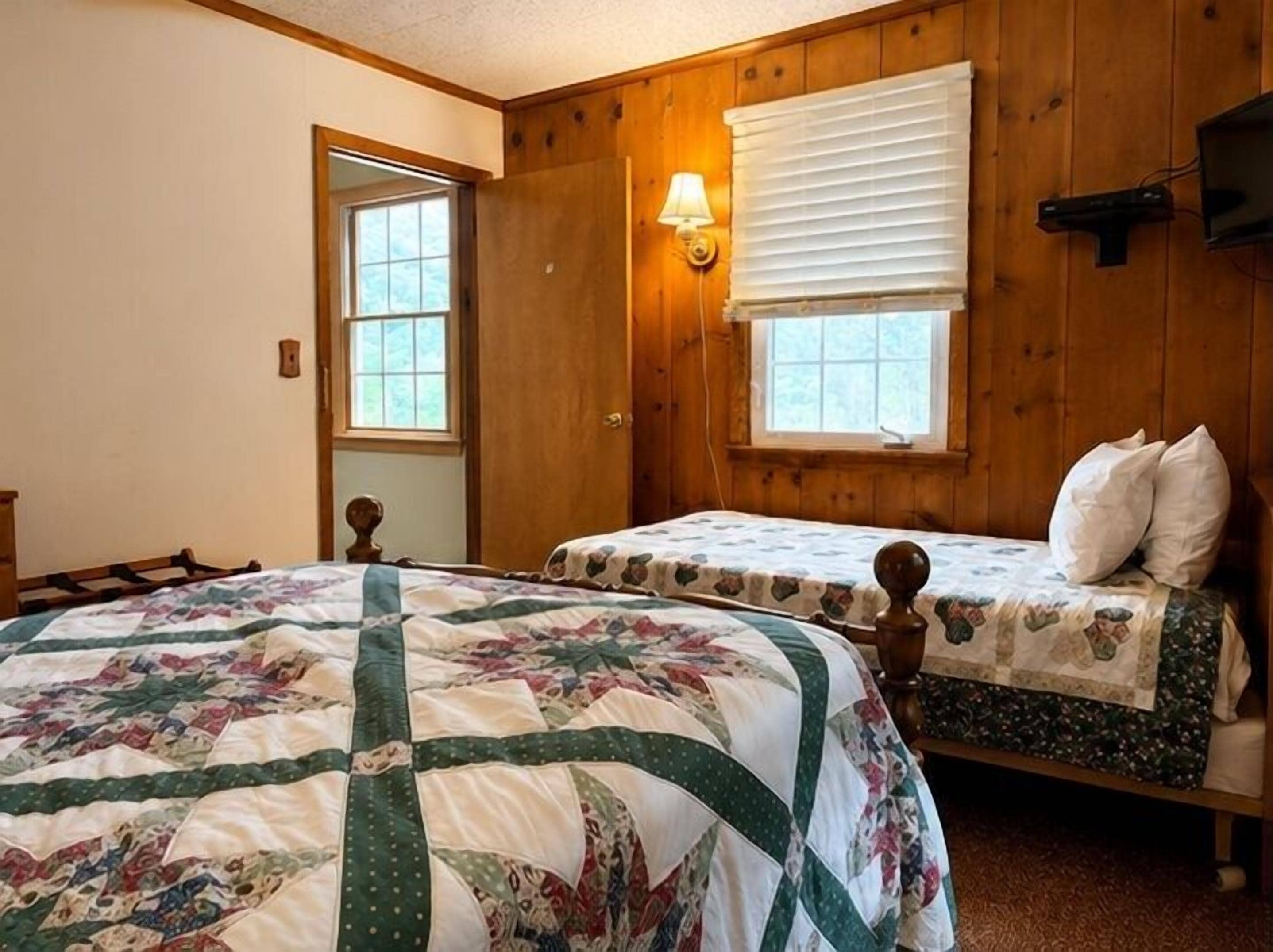 A room with two beds, wooden walls, a window with blinds, and a wall-mounted TV.