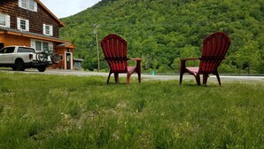 En gräsyta med två röda Adirondack-stolar och en vit lastbil parkerad framför en träbyggnad, med ett berg i bakgrunden.