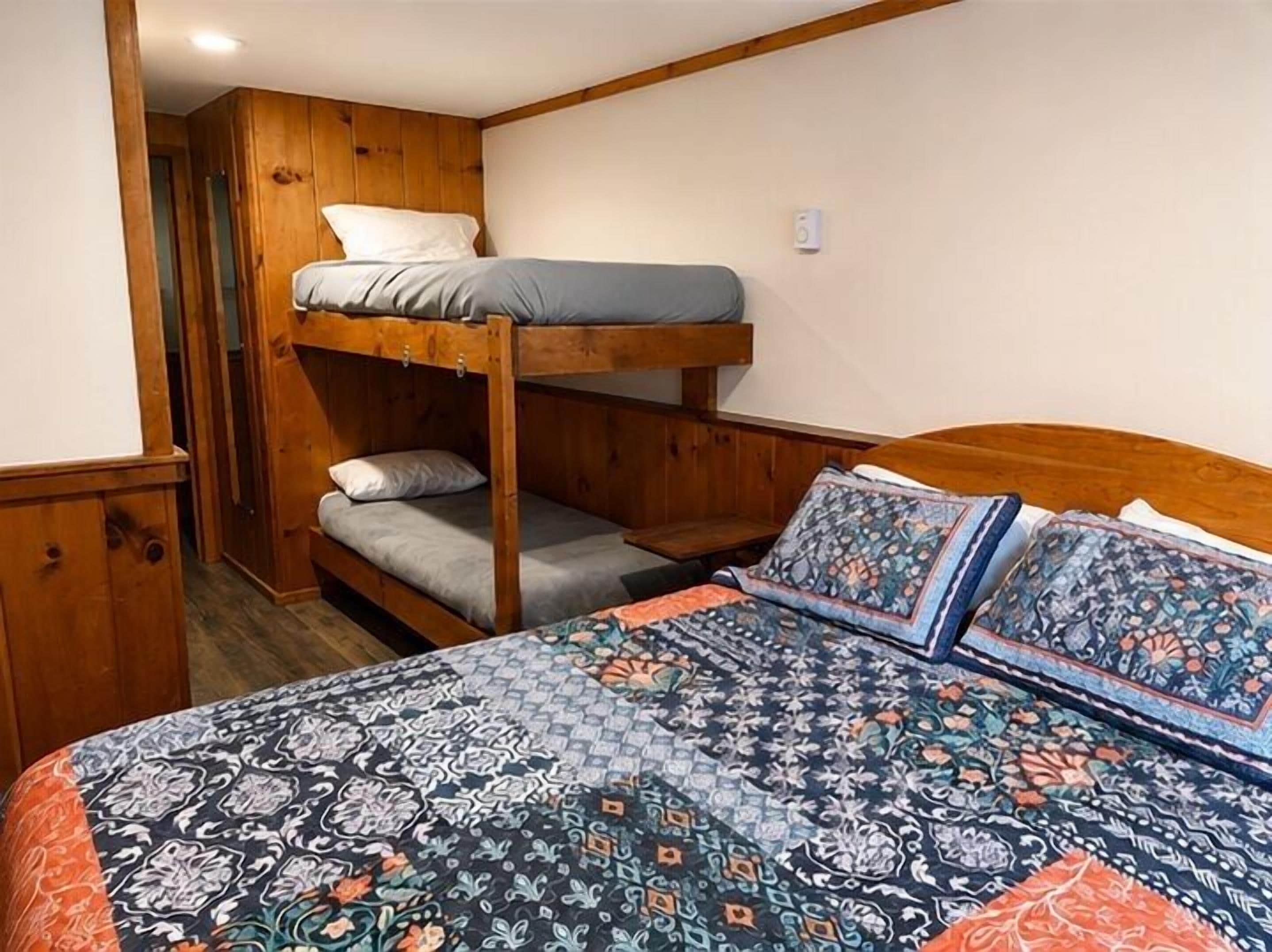 A bunk bed with a patterned bedspread and pillows, a wooden wardrobe, and a door leading to another room.