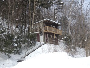 A cabin with a wooden deck and a staircase leading up to it, surrounded by trees and snow. - Turn of River Lodge (Killington)