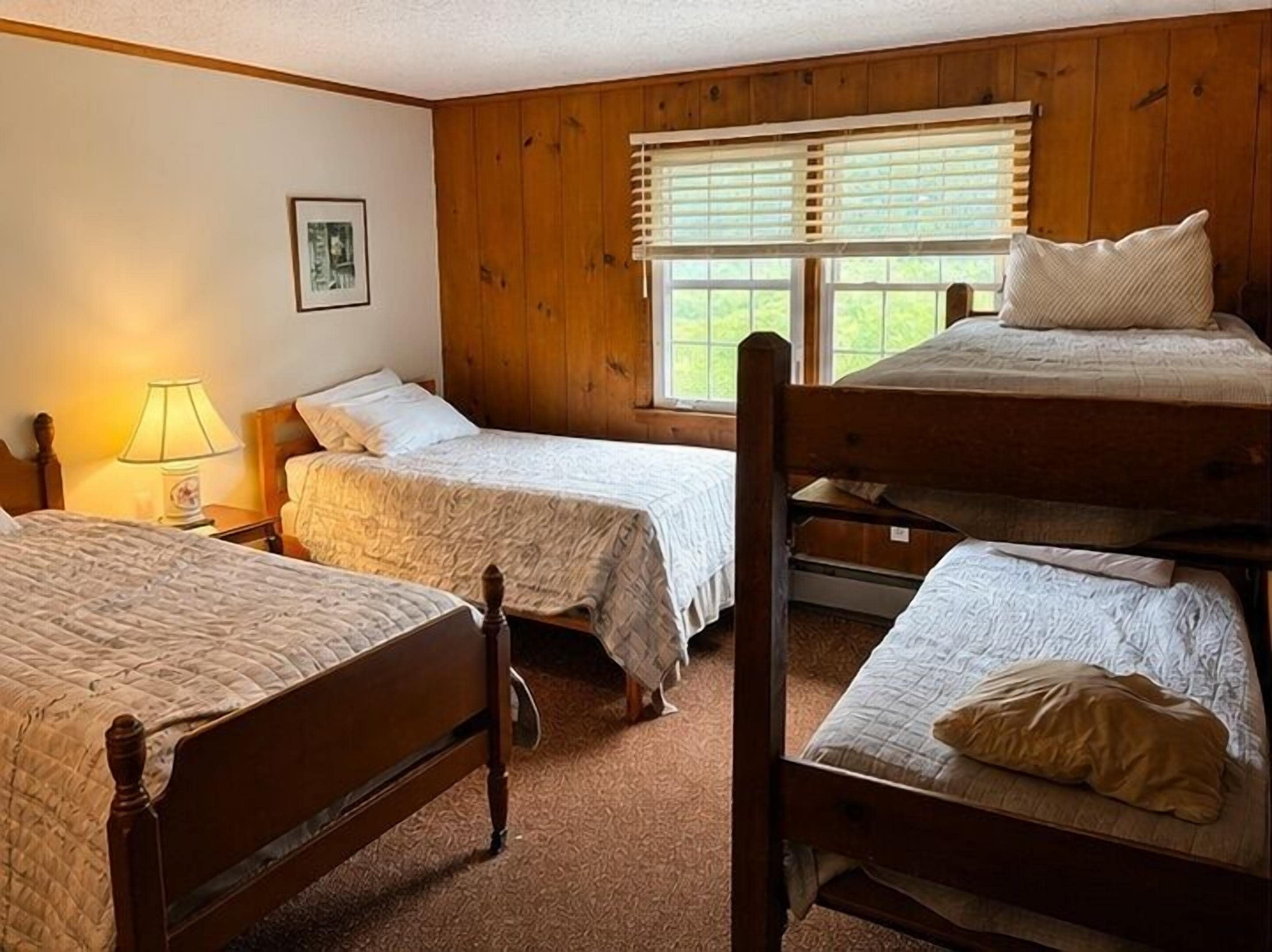 A room with three bunk beds, wooden walls, a window with blinds, and a single bedside table with a lamp.