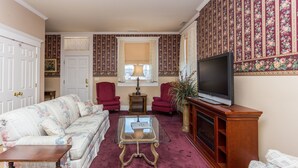 Ein Wohnzimmer mit einem floralen Sofa, zwei roten Sesseln, einem Glastisch, einer Holz-TV-Konsole und einem großen Fenster mit Vorhängen.