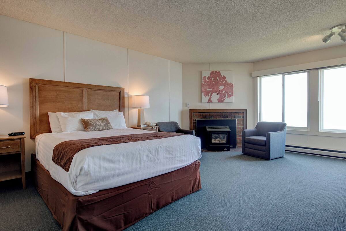A hotel room with a large bed, a fireplace, a chair, and a large window.