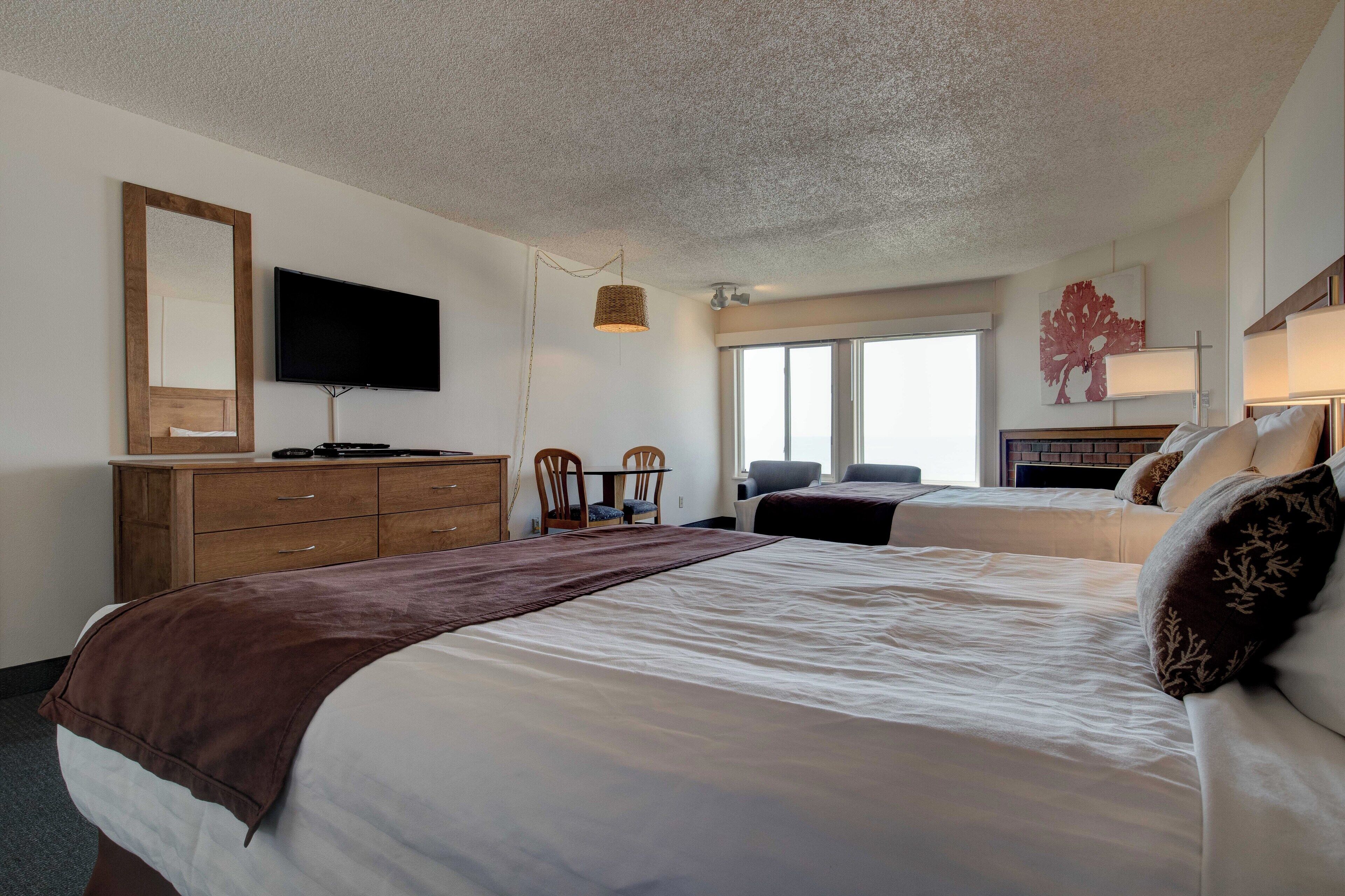 A hotel room with a large bed, a wooden dresser, a flat-screen TV, a chair, a small table, and a window with a view.
