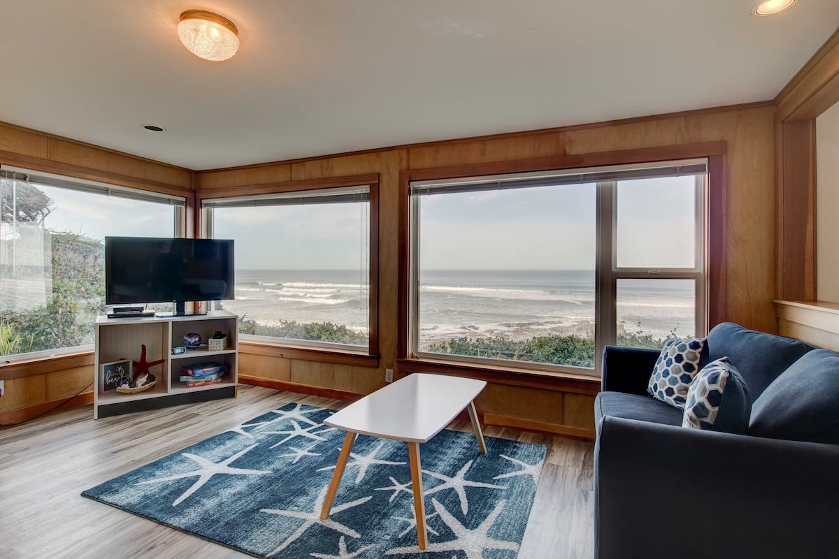 A living room with a TV, a small table, and a sofa with a view of the beach.