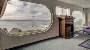 Een kamer met een groot ovaal raam dat uitzicht biedt op de oceaan en een elektriciteitsmast. Er is een open haard met een schouw en een stoel.