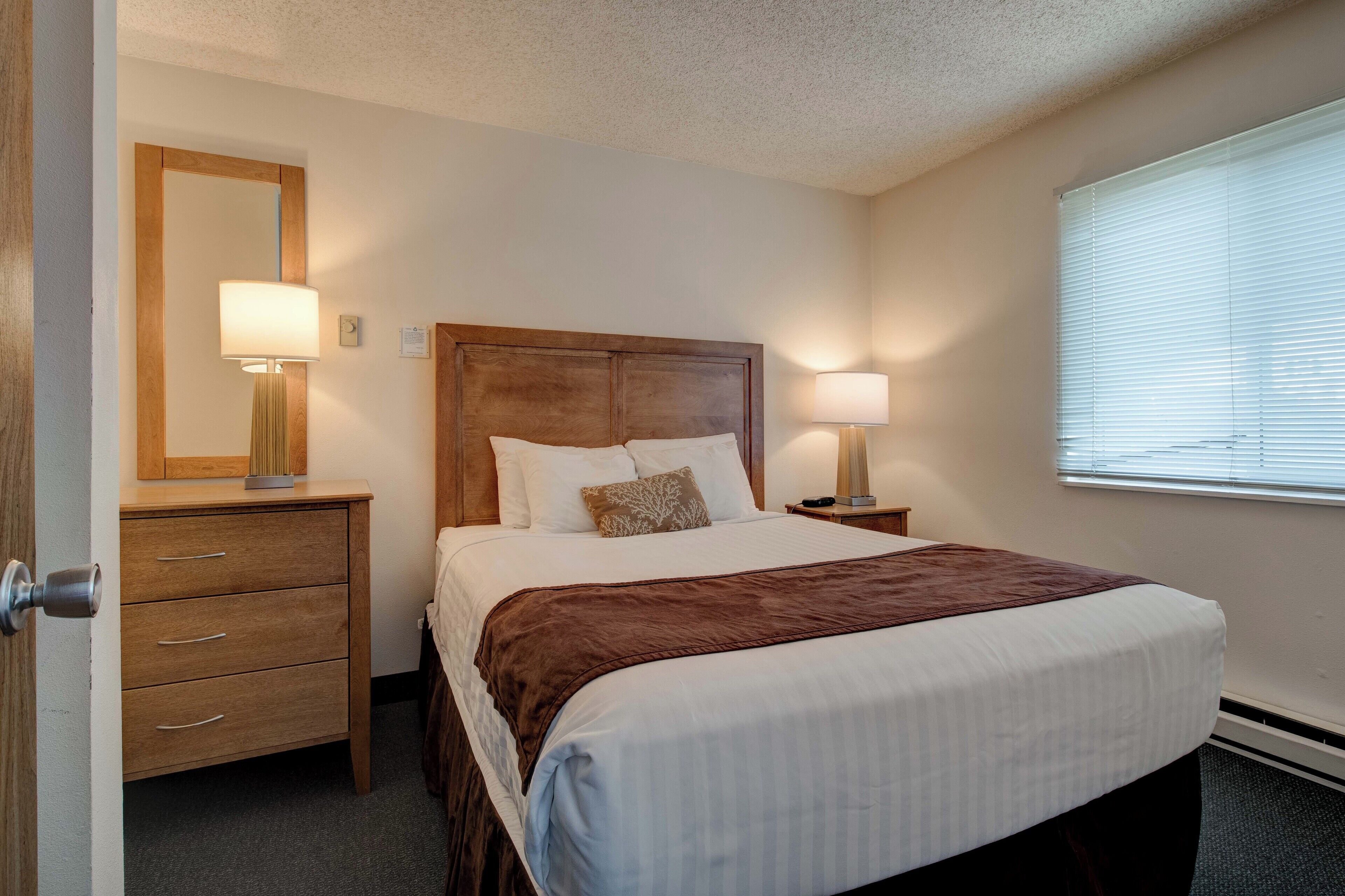 A hotel room with a wooden headboard, a large bed with white bedding, two bedside lamps, a wooden nightstand, a mirror, and a window with blinds.