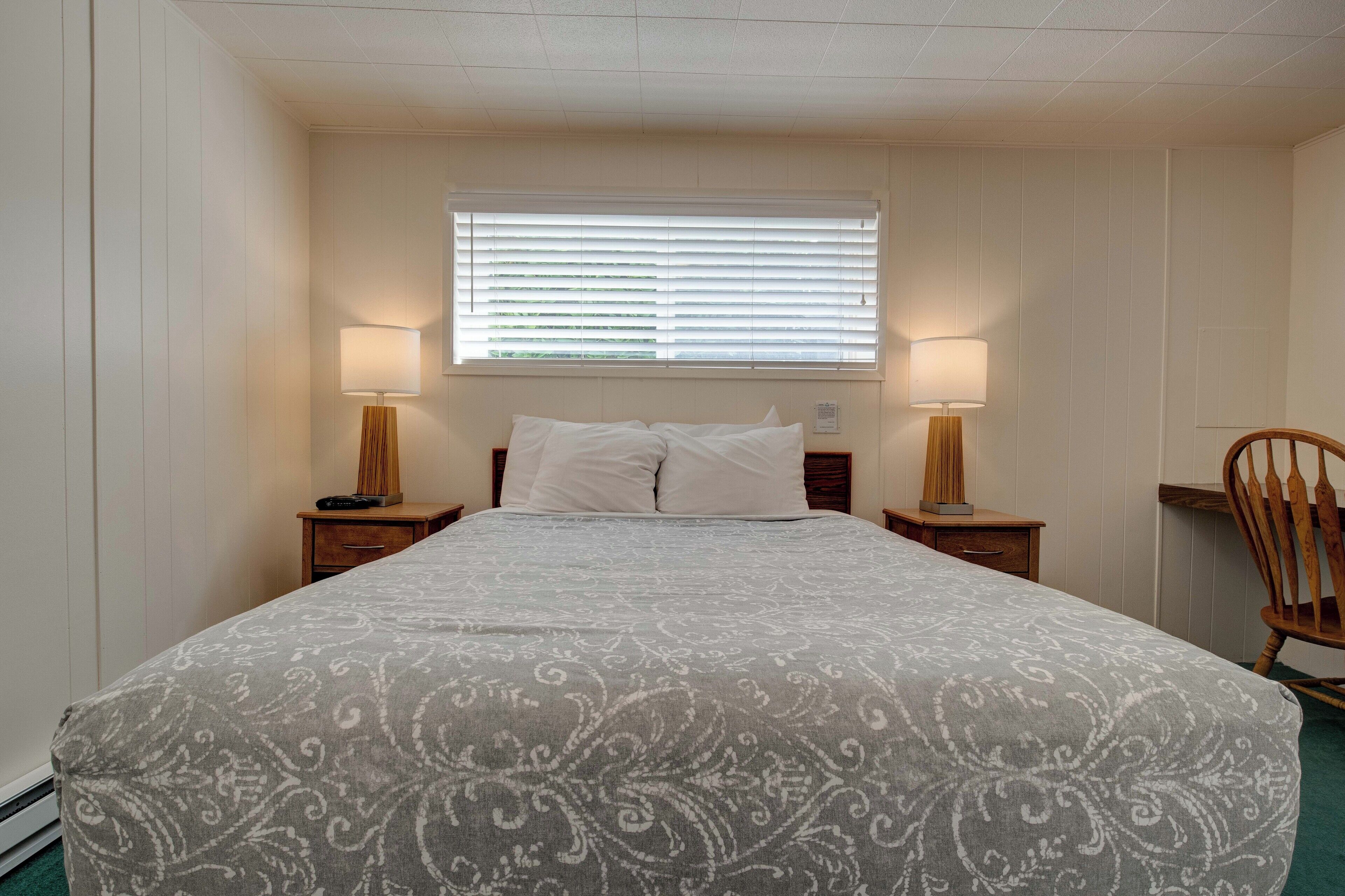 Un dormitorio ordenado con una cama grande, dos mesitas de noche con lámparas, una ventana con persianas y una silla de madera.