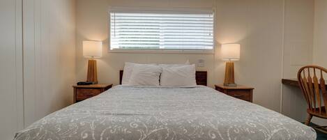 Un dormitorio ordenado con una cama grande, dos mesitas de noche con lámparas, una ventana con persianas y una silla de madera.