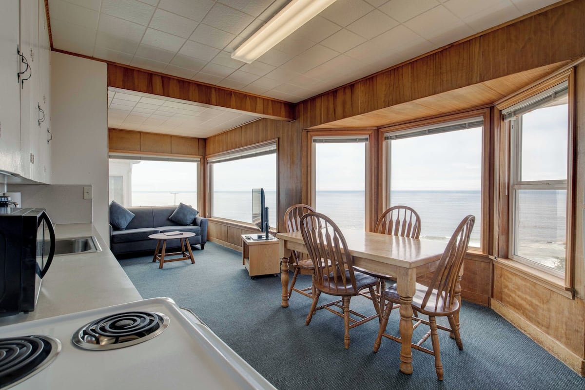 A compact kitchen with a stove, a dining table with chairs, a sofa, and large windows offering a view of the sea.