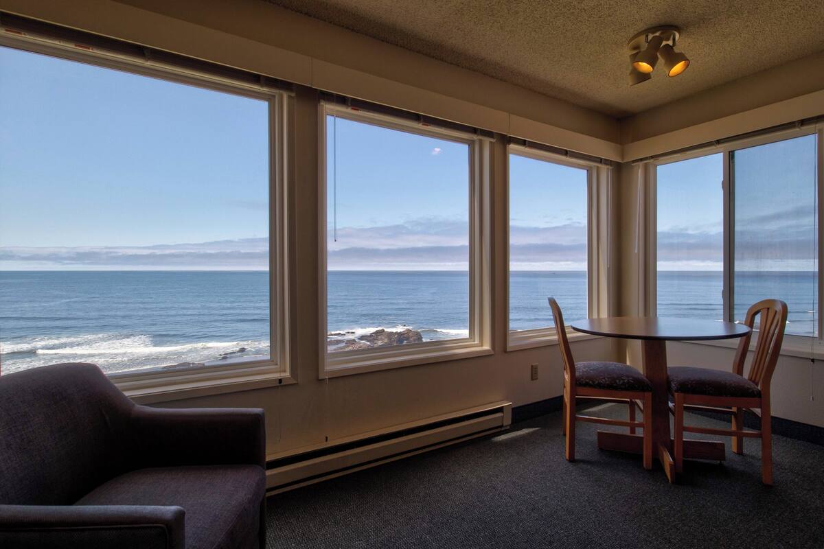 A room with large windows overlooking the ocean, a table with chairs, and a heater under the windows.