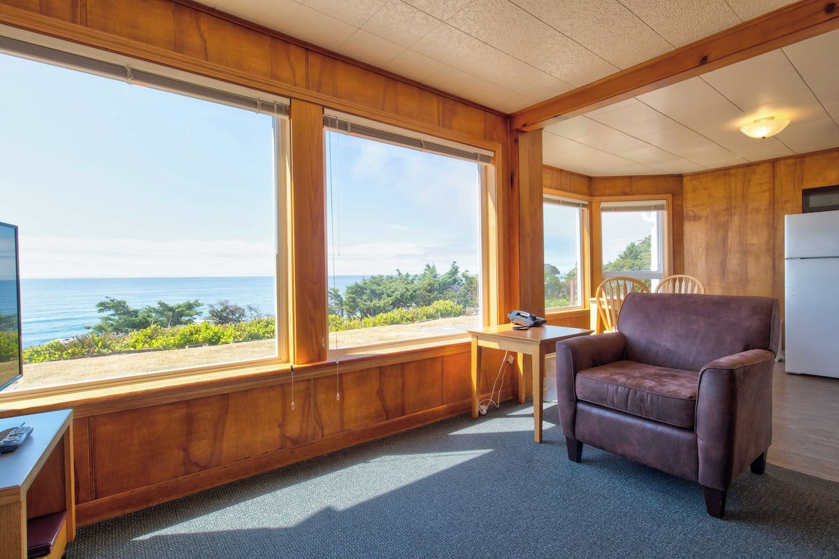 A room with large windows, a brown leather chair, a small table, and a white refrigerator.
