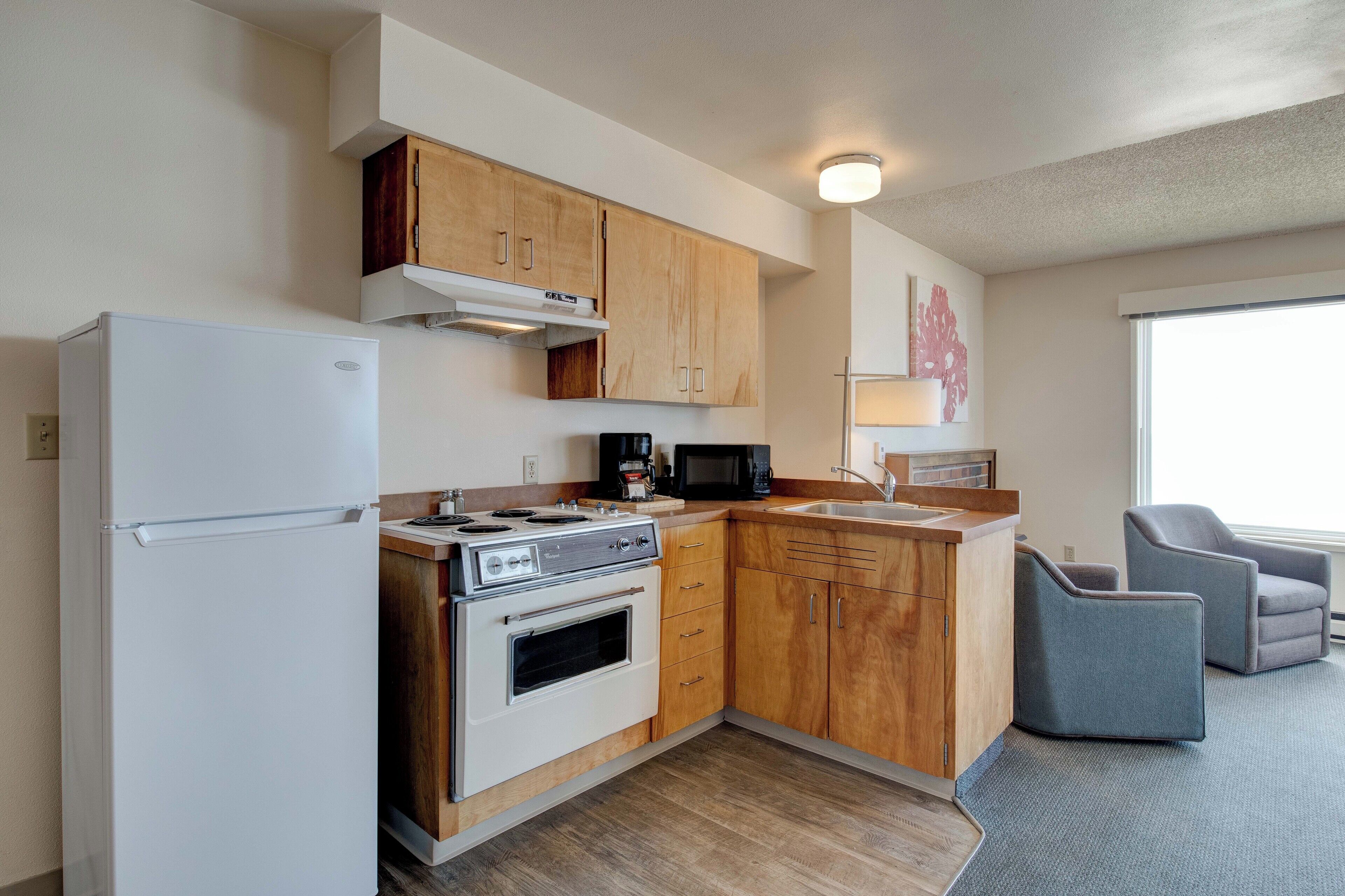 A compact kitchen with wooden cabinets, a white refrigerator, a stove, and a microwave. There is a sink and two chairs in the adjacent living area.